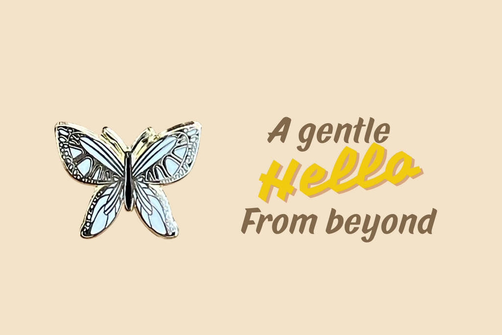 White butterfly enamel pin on beige background with text ‘A gentle hello from beyond’ symbolizing peace and remembrance.